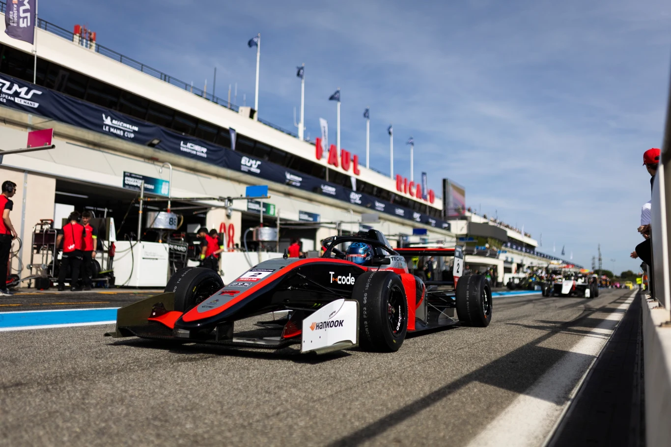 Formuła wyścigowy samochód z numerem 8 ustawiony na torze przy pitstopie, w tle zespół techniczny oraz zaplecze toru wyścigowego.