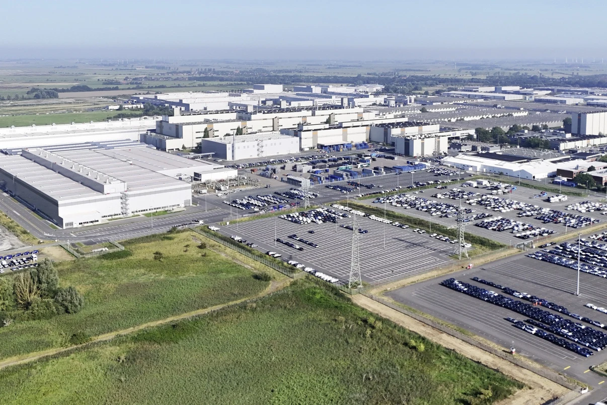 Rozległy kompleks przemysłowy z dużymi halami produkcyjnymi i magazynami, otoczony licznymi parkingami z zaparkowanymi samochodami oraz siecią dróg dojazdowych, sąsiadujący z terenami zielonymi.