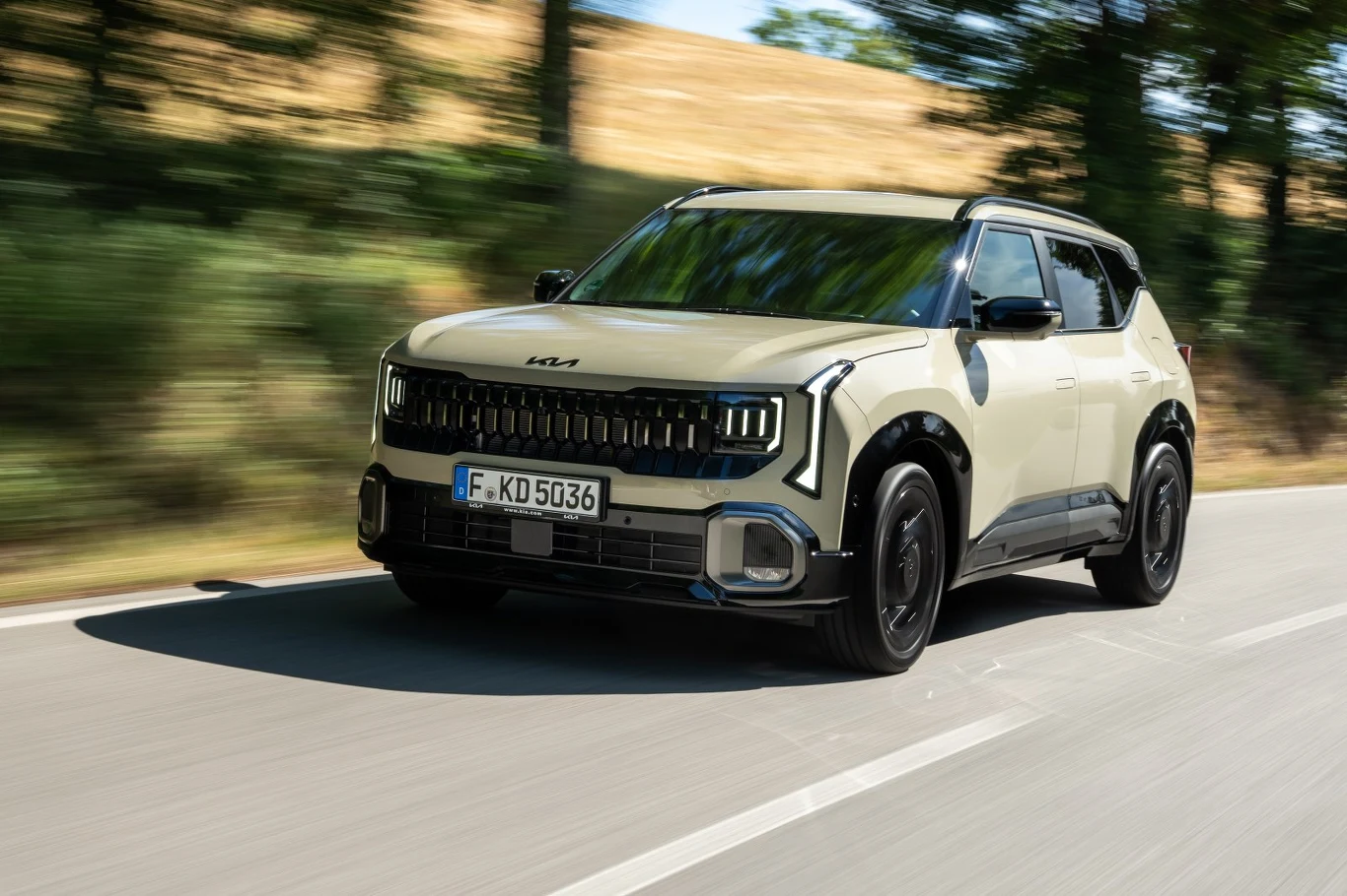 Jasny SUV marki Kia z nowoczesnym designem reflektorów i czarnymi detalami porusza się dynamicznie po jednopasmowej drodze, otoczonej rozmytą zielenią drzew i polami.