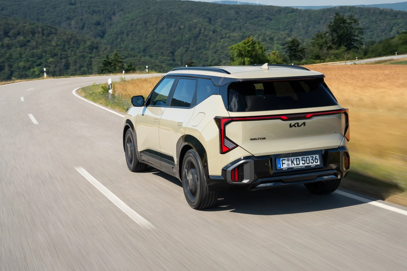 Jasny SUV marki Kia z nowoczesnym designem i czarnymi akcentami porusza się drogą prowadzącą przez zielone wzgórza.