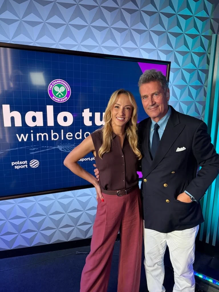 Tomasz Tomaszewski i Paula Kania Kobieta i mężczyzna stoją razem przed ekranem z logo Wimbledonu oraz napisem 'halo tu wimbledon', uśmiechając się do kamery w studiu telewizyjnym Polsat Sport.