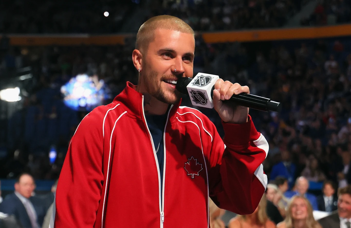 Justin Bieber Mężczyzna w czerwonej kurtce z kanadyjskim liściem klonowym trzyma mikrofon i przemawia do publiczności na dużej arenie.