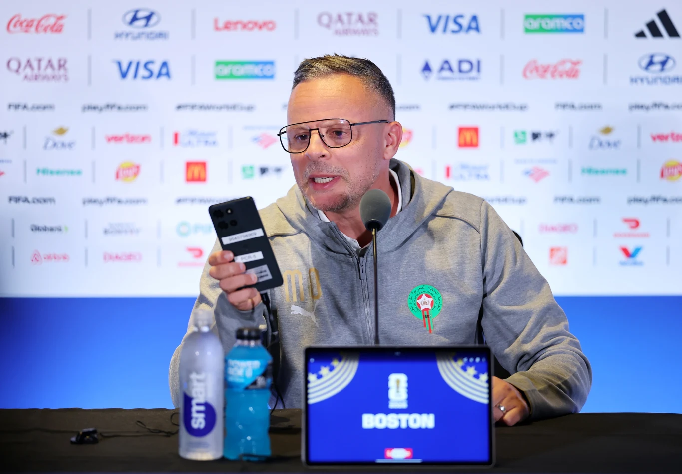 Mohamed Ouahbi Mężczyzna w okularach w szarej bluzie z emblematem, siedzący przy stole konferencyjnym, trzyma telefon przed mikrofonem, na stole przed nim laptop z napisem Boston oraz dwie butelki napojów; tło wypełniają logotypy sponsorów.