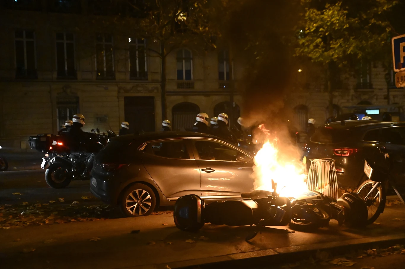 Płonący skuter leżący na chodniku obok zaparkowanego samochodu, w tle grupa policjantów w kaskach stojących na ulicy nocą.