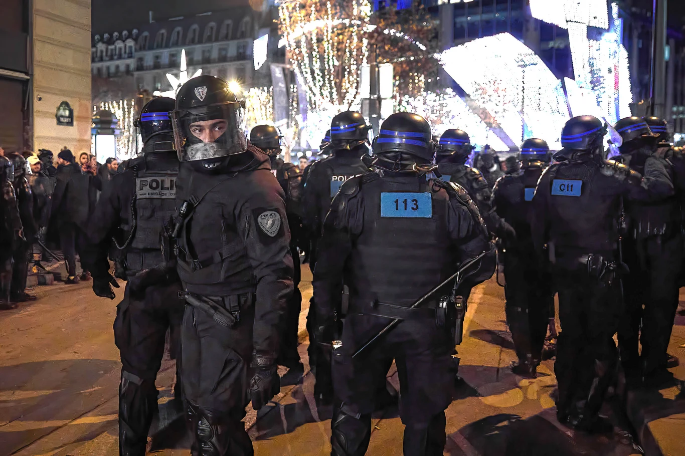 Grupa policjantów w pełnym umundurowaniu ochronnym i hełmach kordonuje ulicę w miejskiej przestrzeni nocą, na drugim planie widoczne świąteczne oświetlenie i zgromadzeni ludzie.