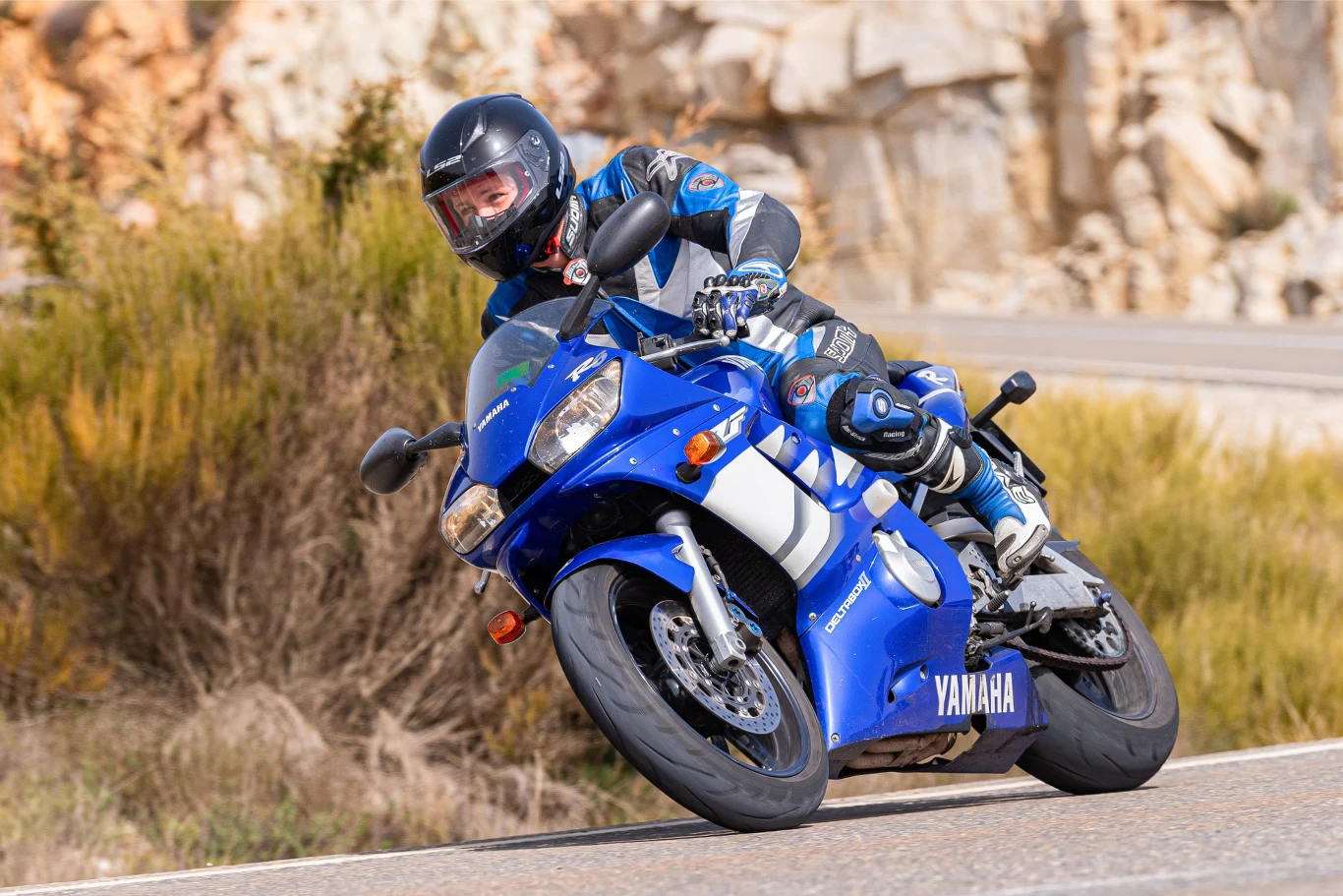 Motocyklista w profesjonalnym kombinezonie pokonuje zakręt na sportowym, niebieskim motocyklu Yamaha jadąc dynamicznie po asfaltowej drodze.