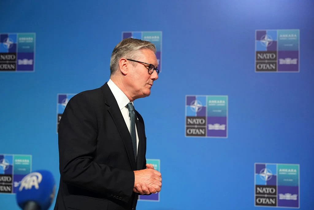Mężczyzna w garniturze i okularach idący obok ściany z logo NATO oraz mikrofonem na pierwszym planie.