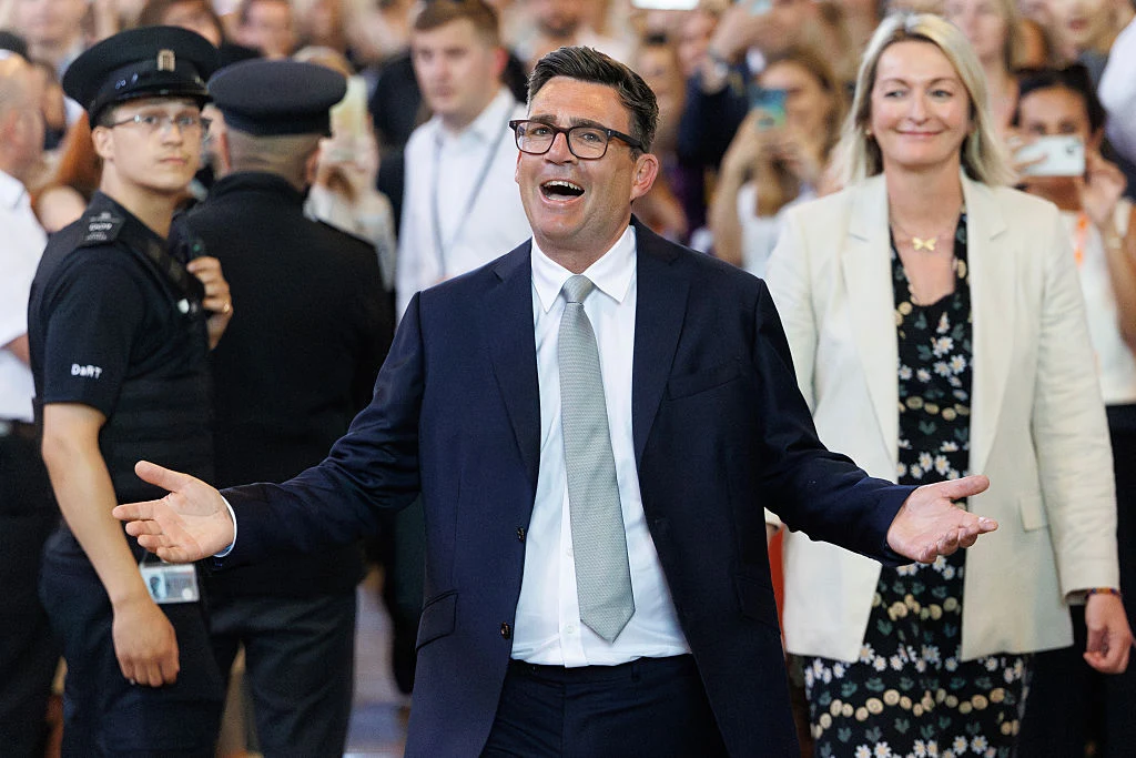 Uśmiechnięty mężczyzna w garniturze z rozłożonymi rękami stoi na pierwszym planie, w tle kobieta w jasnym żakiecie i tłum ludzi, wśród których widoczny jest policjant.
