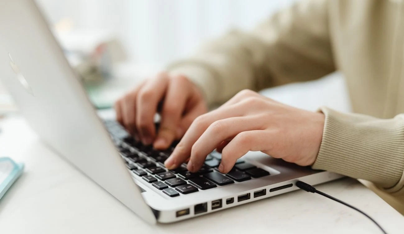 Absolwenci niezmiennie najlepiej radzą sobie z językiem angielskim Osoba w jasnej bluzie pisząca na srebrnym laptopie z czarnymi klawiszami. Laptop stoi na jasnym biurku.