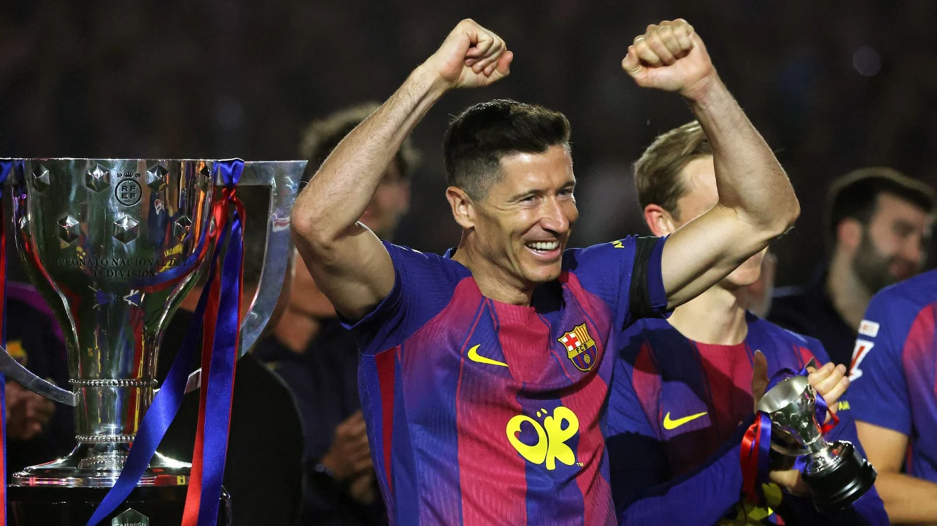 Piłkarz drużyny FC Barcelona w koszulce klubowej z uniesionymi rękami w geście triumfu, stojący obok dużego trofeum ozdobionego niebiesko-czerwonymi wstążkami.