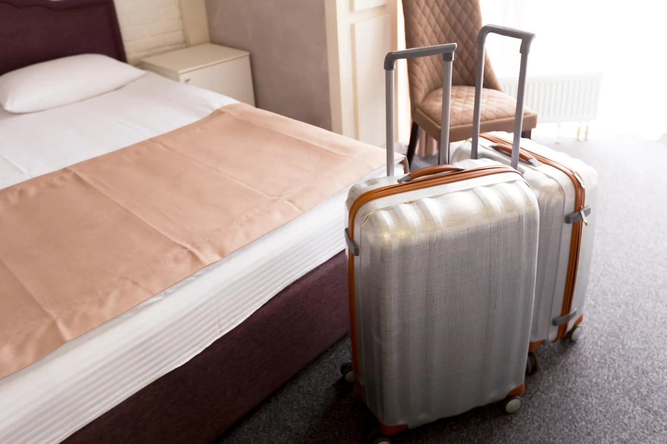 Dlaczego w hotelu warto wstawić walizkę pod prysznic? Dwie srebrne walizki na kółkach stoją obok łóżka w schludnym pokoju hotelowym z krzesłem przy łóżku.