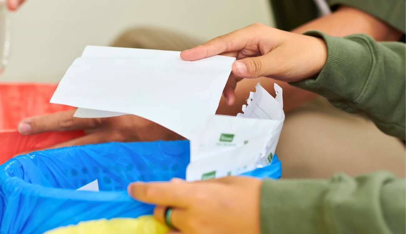 Gdzie wyrzucać papier kredowy? Dłonie wyrzucają papier kredowy do niebieskiego pojemnika na odpady papierowe.