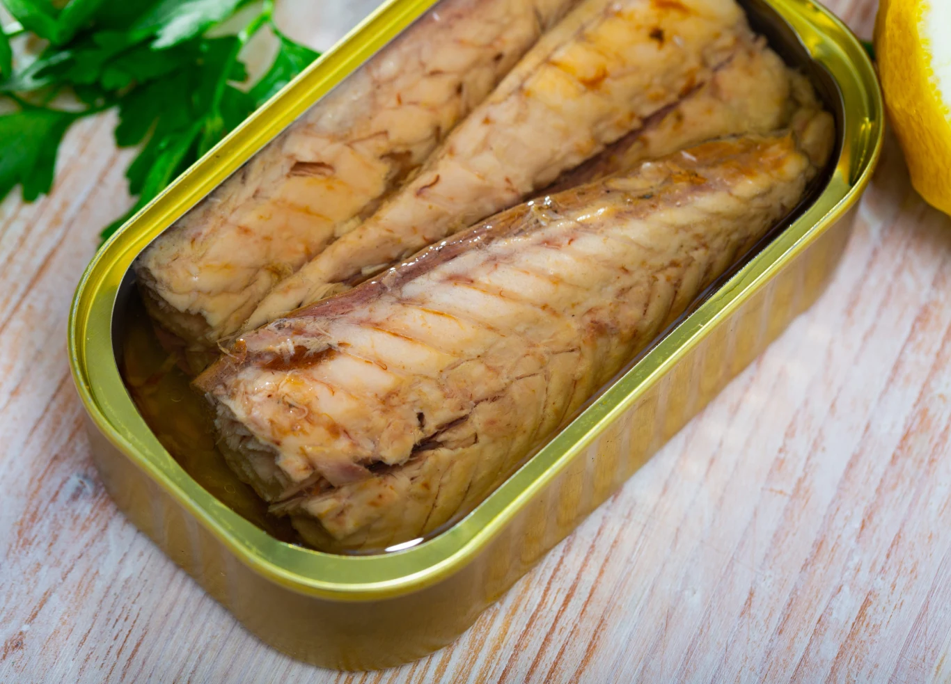 Makrela z puszki może być dobrą przekąską, która dostarcza cennych kwasów omega-3 i białka. Trzy kawałki makreli w oleju w otwartej puszce na drewnianej powierzchni z natką pietruszki i cytryną.