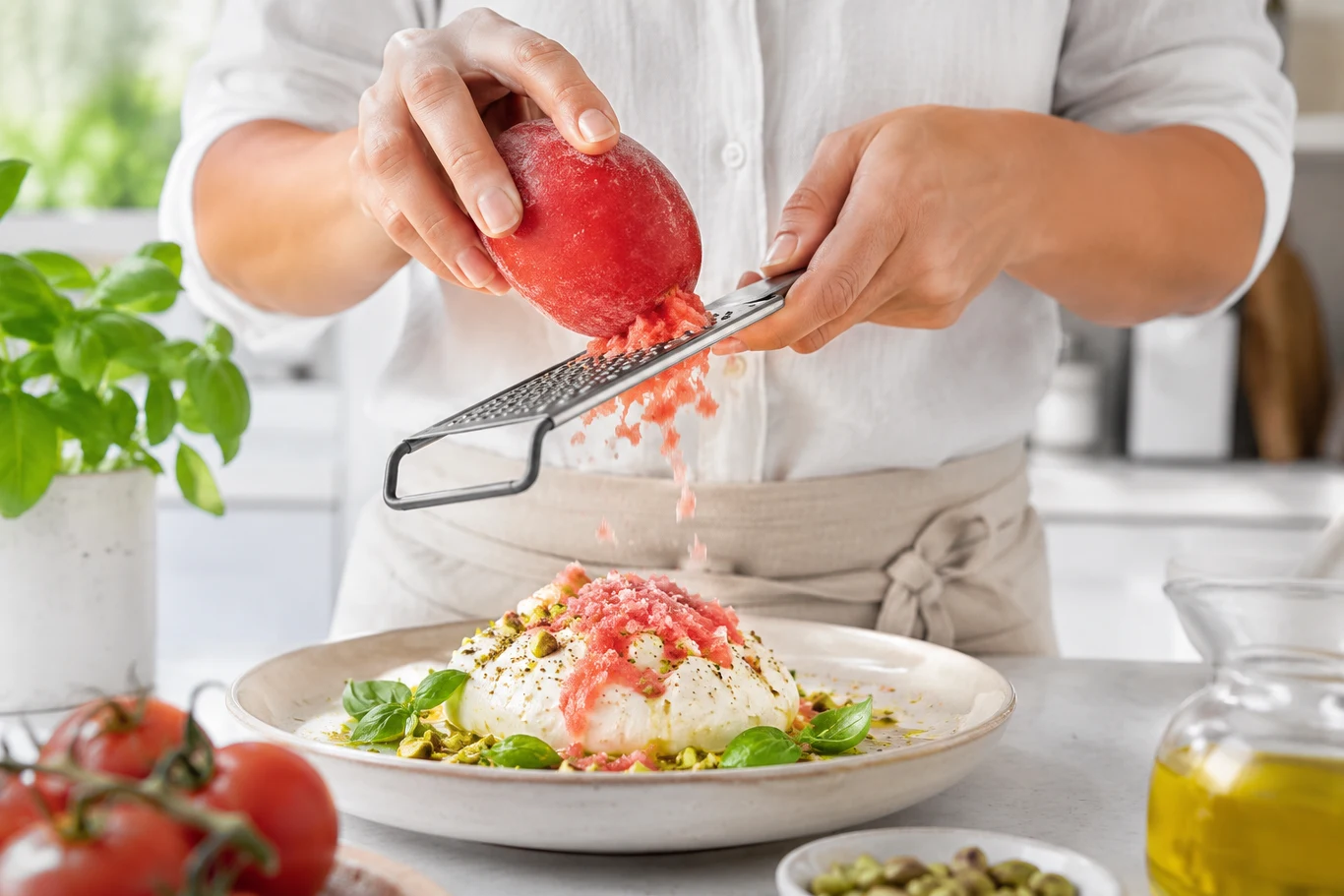 Personka ściera dojrzałego pomidora na tarce nad talerzem z serem burrata, pistacjami i świeżą bazylią.