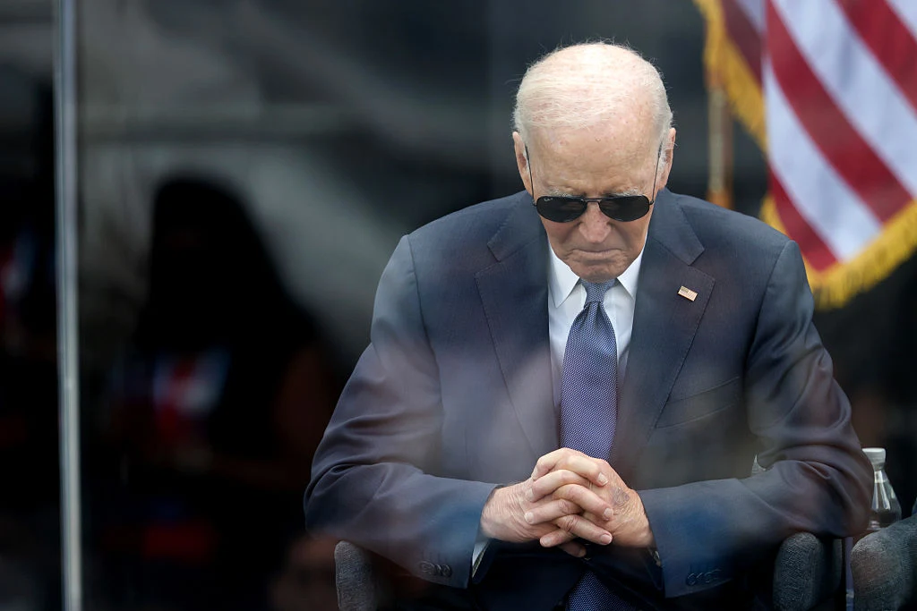 Były prezydent USA Joe Biden odszedł ze stanowiska w wieku 82 lat Mężczyzna w garniturze i okularach przeciwsłonecznych siedzi z pochyloną głową i splecionymi dłońmi, w tle flaga Stanów Zjednoczonych.