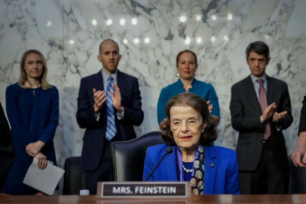 Senator z Kalifornii Dianne Feinstain zmarła w wieku 90 lat, pełniąc nadal funkcję Starsza kobieta siedzi przy stole z tabliczką „Mrs. Feinstein”, z poważnym wyrazem twarzy, za nią stoi czworo ludzi w eleganckich ubraniach, niektórzy klaszczą.