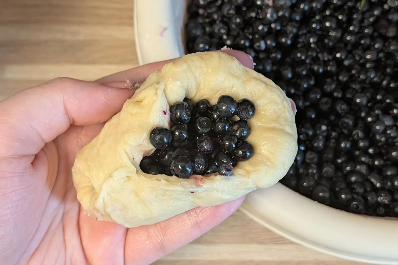 Brzegi bułeczki zlep dokładnie, tak, aby nie rozkleiły się one podczas pieczenia Dłoń trzyma kawałek rozwałkowanego ciasta wypełnionego świeżymi jagodami, obok miska pełna owoców.