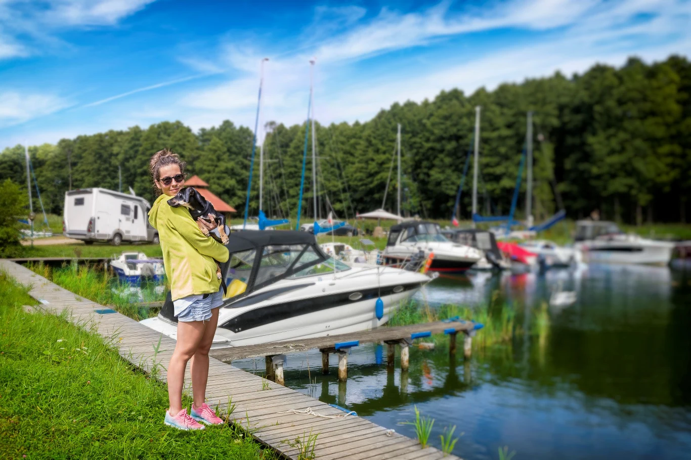 Kobieta w jasnej kurtce i różowych butach stoi na drewnianym pomoście, trzymając psa na rękach; za nią zacumowane jachty i łódki przy zielonym brzegu, w tle las oraz kamper.