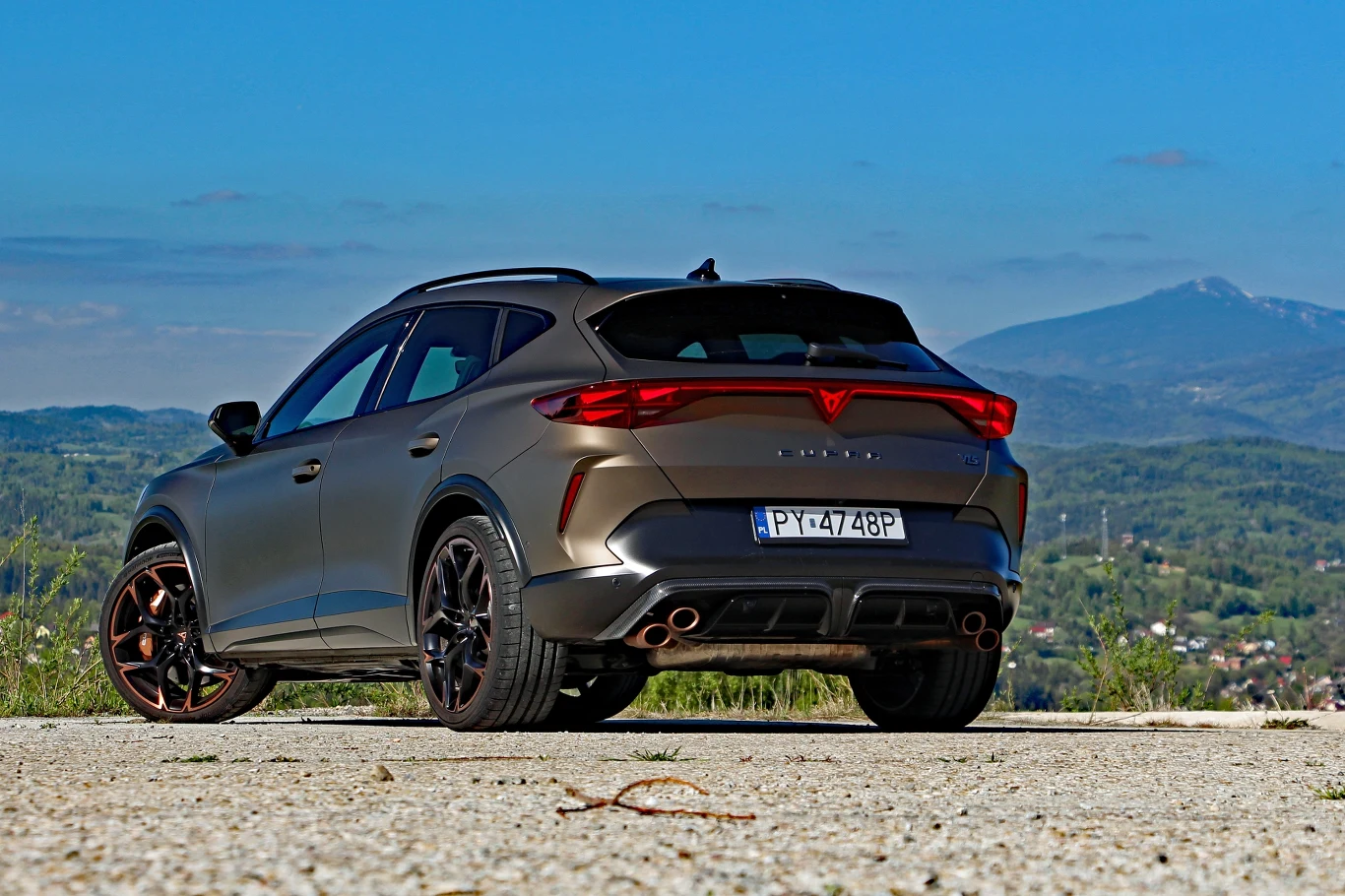 Sportowy SUV w matowym szarym kolorze z agresywną linią tylnych świateł i czterema końcówkami wydechu stoi na utwardzonym podłożu z widokiem na zielone wzgórza oraz górski szczyt w oddali.