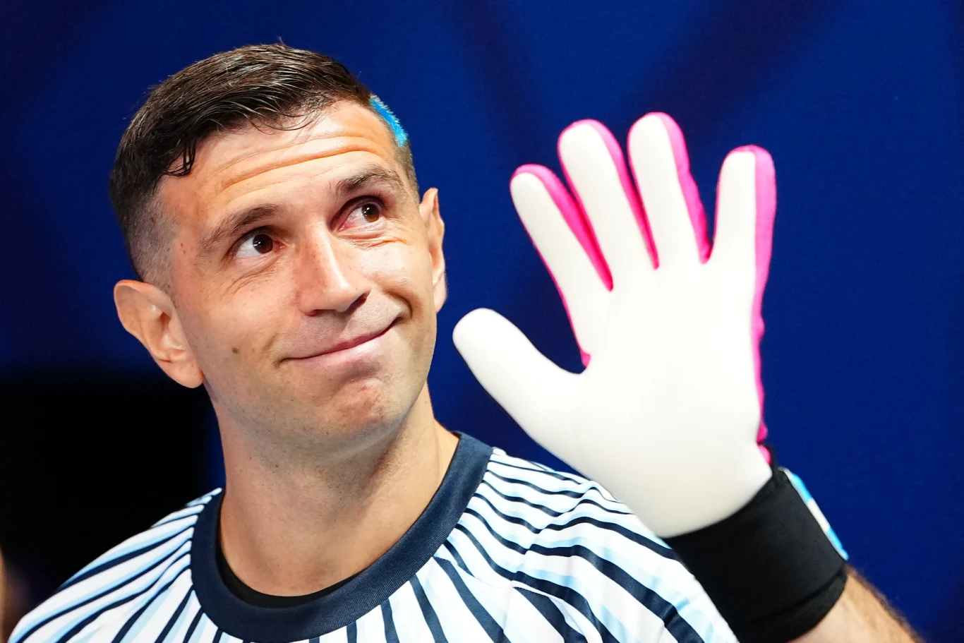 Mężczyzna w pasiastym sportowym stroju unosi rękę w biało-różowej rękawicy bramkarskiej, z łagodnym uśmiechem na twarzy spogląda w bok na tle niebieskiej ściany.