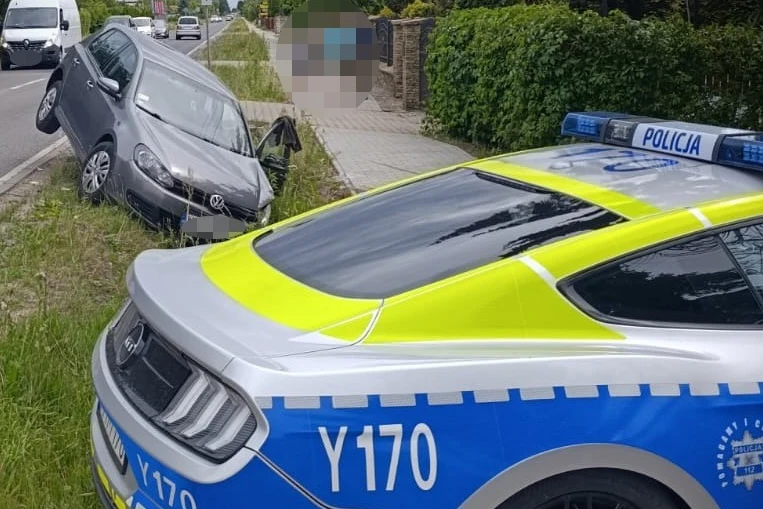 Samochód osobowy wjechał na pobocze i utknął w rowie, obok stoi radiowóz policji z włączonymi sygnałami świetlnymi.