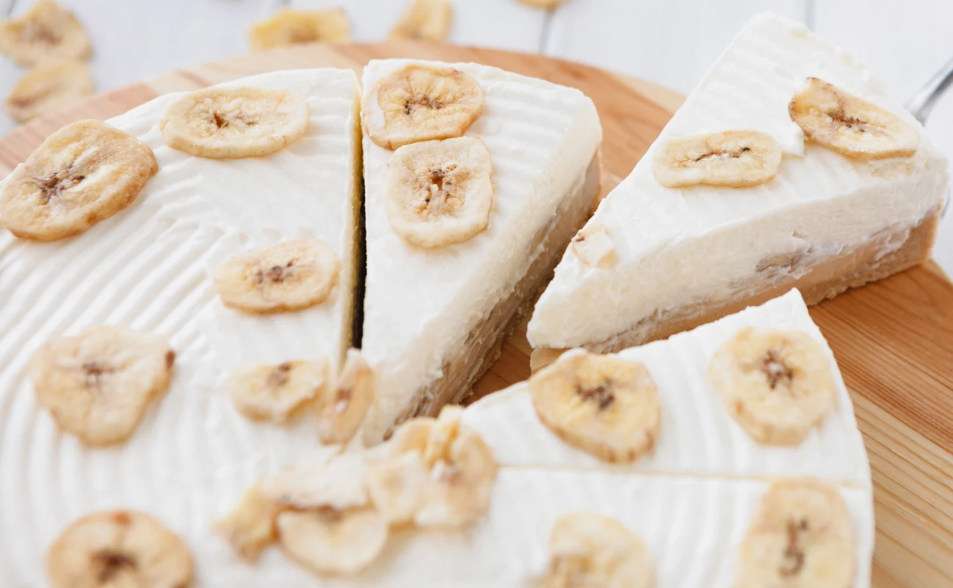 To idealne ciasto na upały. Zrobisz je bez użycia piekarnika Okrągły sernik bananowy bez pieczenia z jasną polewą i kawałkami bananów, pokrojony na porcje.