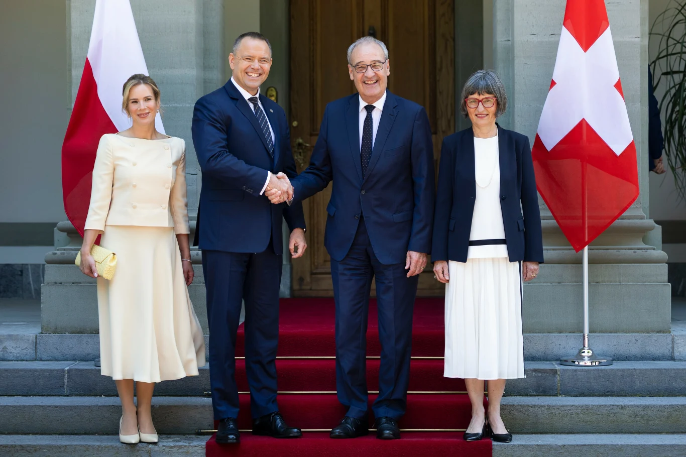 Cztery osoby stojące na czerwonym dywanie przed wejściem do budynku, dwaj mężczyźni w garniturach ściskają sobie dłonie, po bokach stoją dwie kobiety w eleganckich jasnych strojach, po lewej stronie widoczna flaga Polski, po prawej flaga Szwajcarii.
