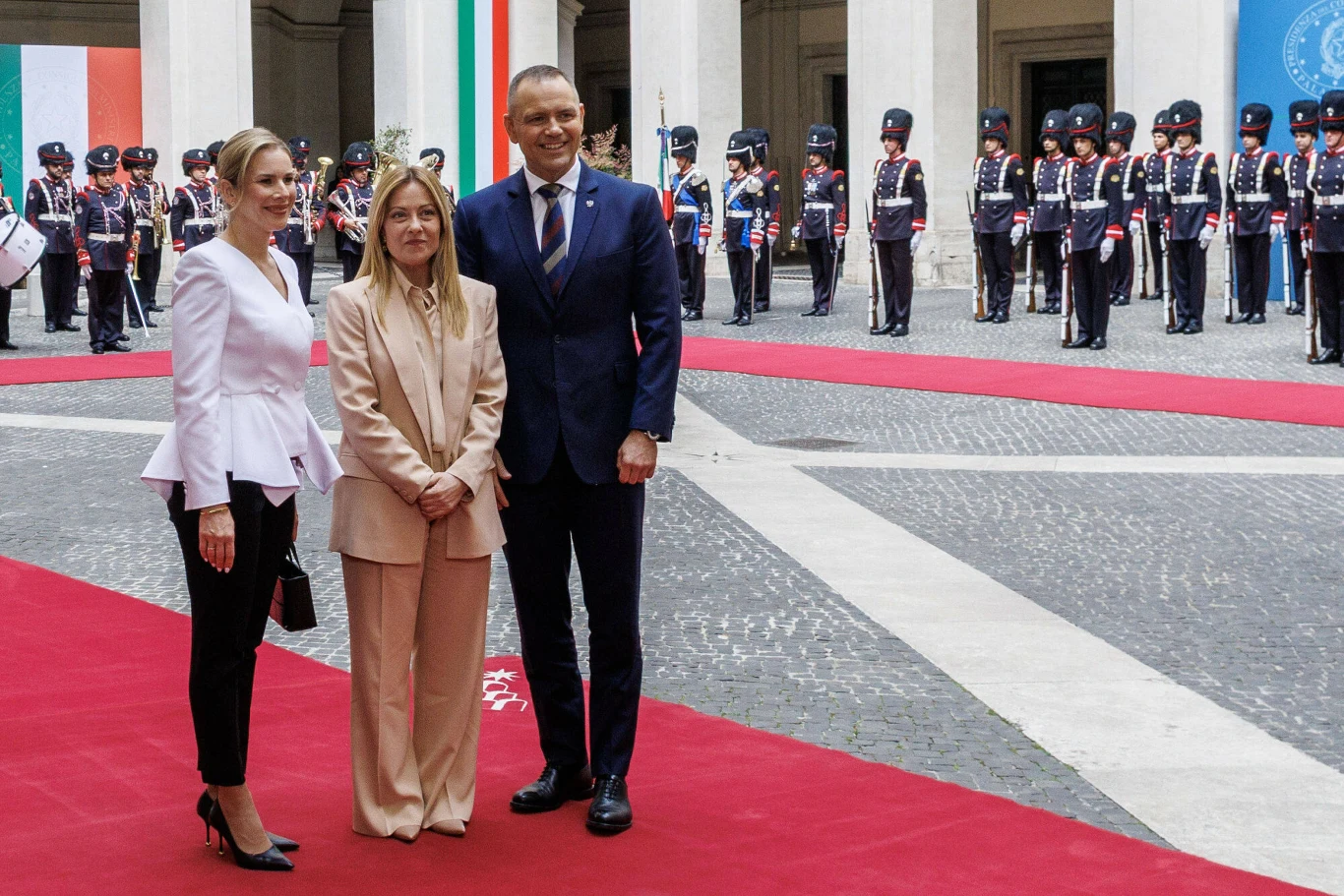 Trzy osoby w eleganckich strojach stoją na czerwonym dywanie, w tle rząd umundurowanych żołnierzy podczas oficjalnej ceremonii.
