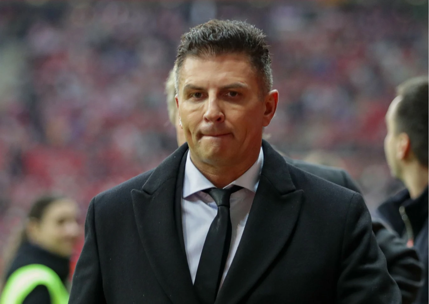Mateusz Borek Mężczyzna w ciemnym płaszczu i białej koszuli z krawatem, stoi na tle zamazanego stadionu i osób w tle.