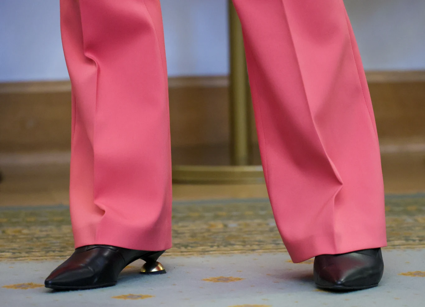 Czarne buty na obcasie z szerokimi różowymi spodniami opadającymi na stopy, ustawione na dywanie z delikatnym wzorem.
