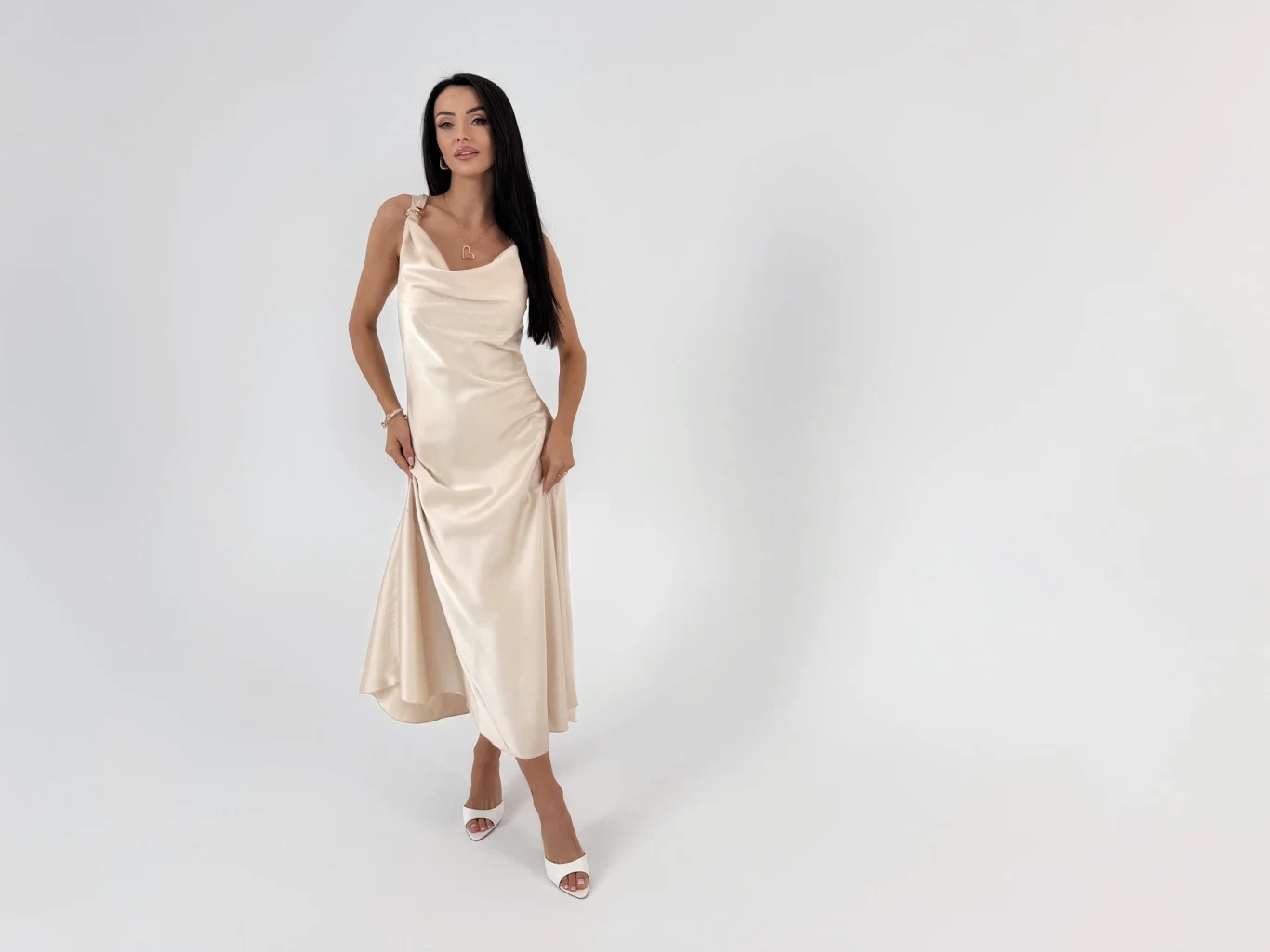 Elegancka beżowa sukienka na wesele na ramiączka marki ModnaKiecka Kobieta z długimi ciemnymi włosami w eleganckiej, kremowej satynowej sukience do połowy łydki i białych sandałach na obcasie na tle jasnej ściany.