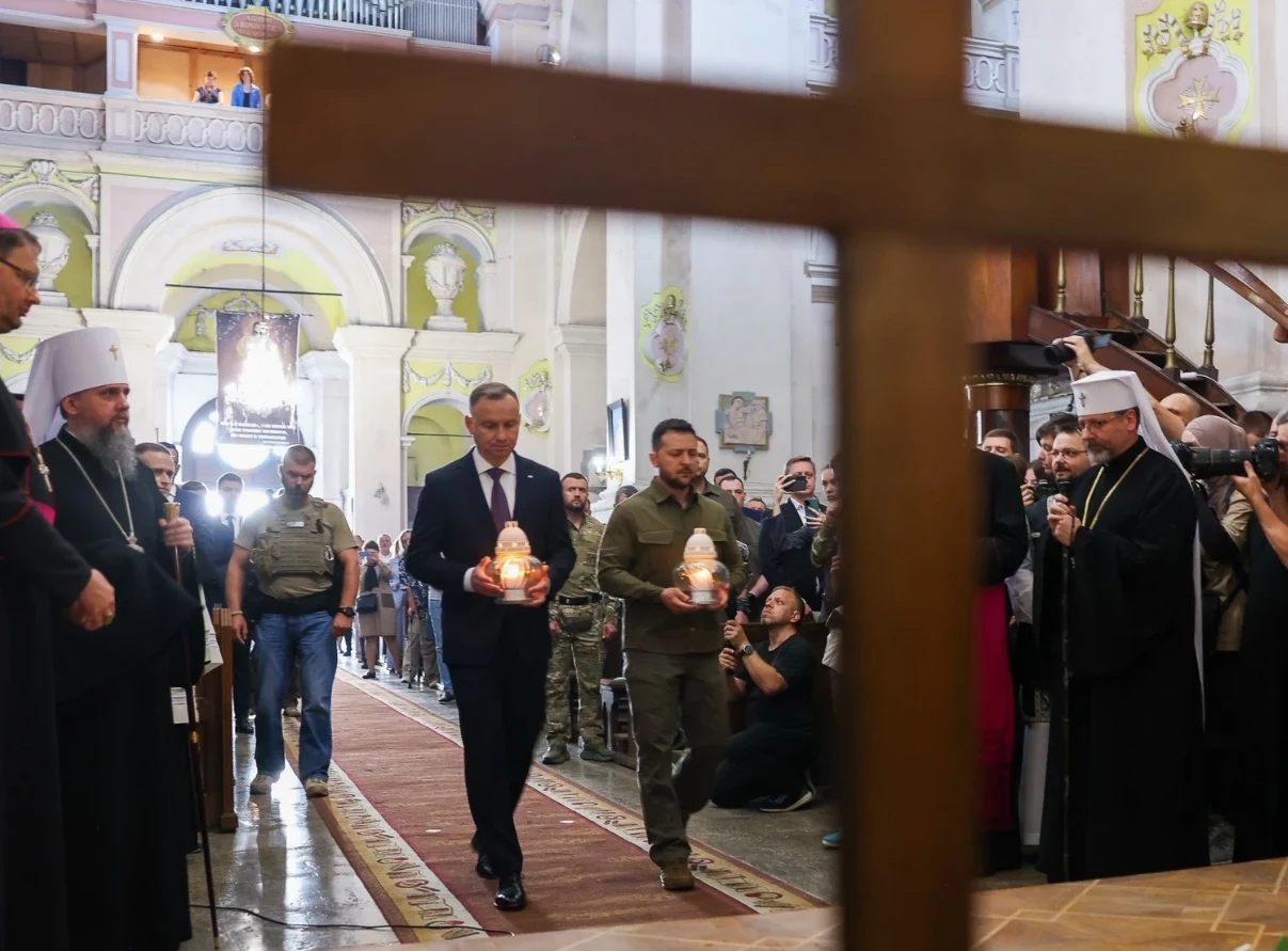 Dwóch mężczyzn niesie zapalone znicze wzdłuż kościoła, otoczonych przez duchownych w szatach liturgicznych oraz zgromadzonych wiernych i fotoreporterów, na pierwszym planie widoczny drewniany krzyż.