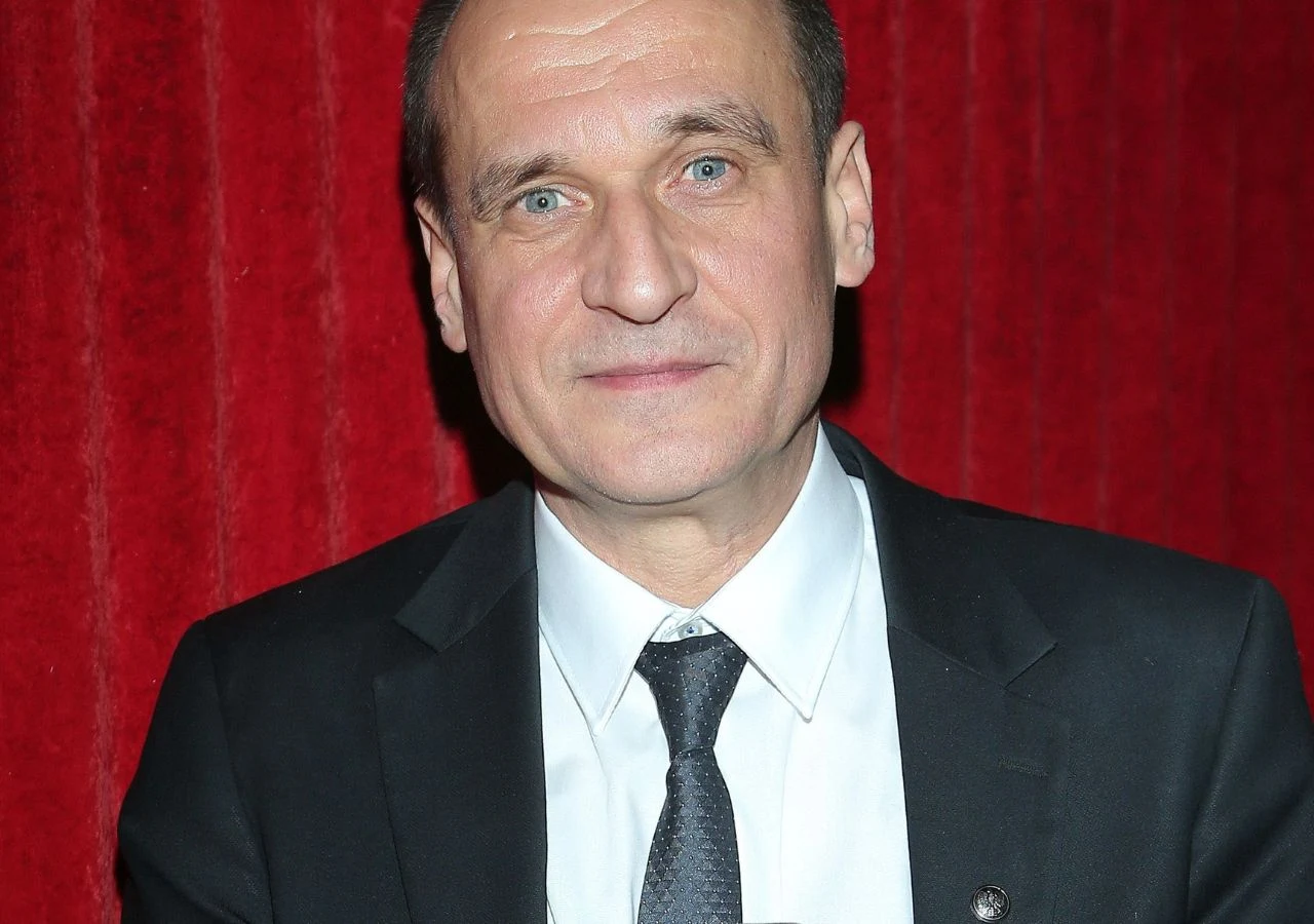 Paweł Kukiz Paweł Kukiz w ciemnym garniturze i białej koszuli, stojący na tle czerwonej kotary.