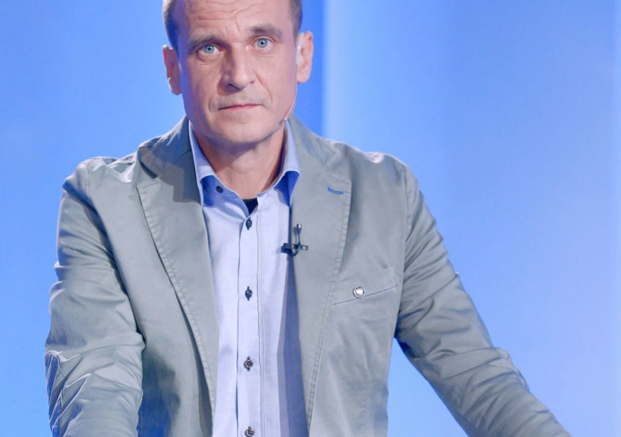 Paweł Kukiz Mężczyzna w jasnej marynarce i błękitnej koszuli stoi przy stole na tle niebieskiego, jednolitego tła, z mikrofonem przypiętym do klapy ubrania.