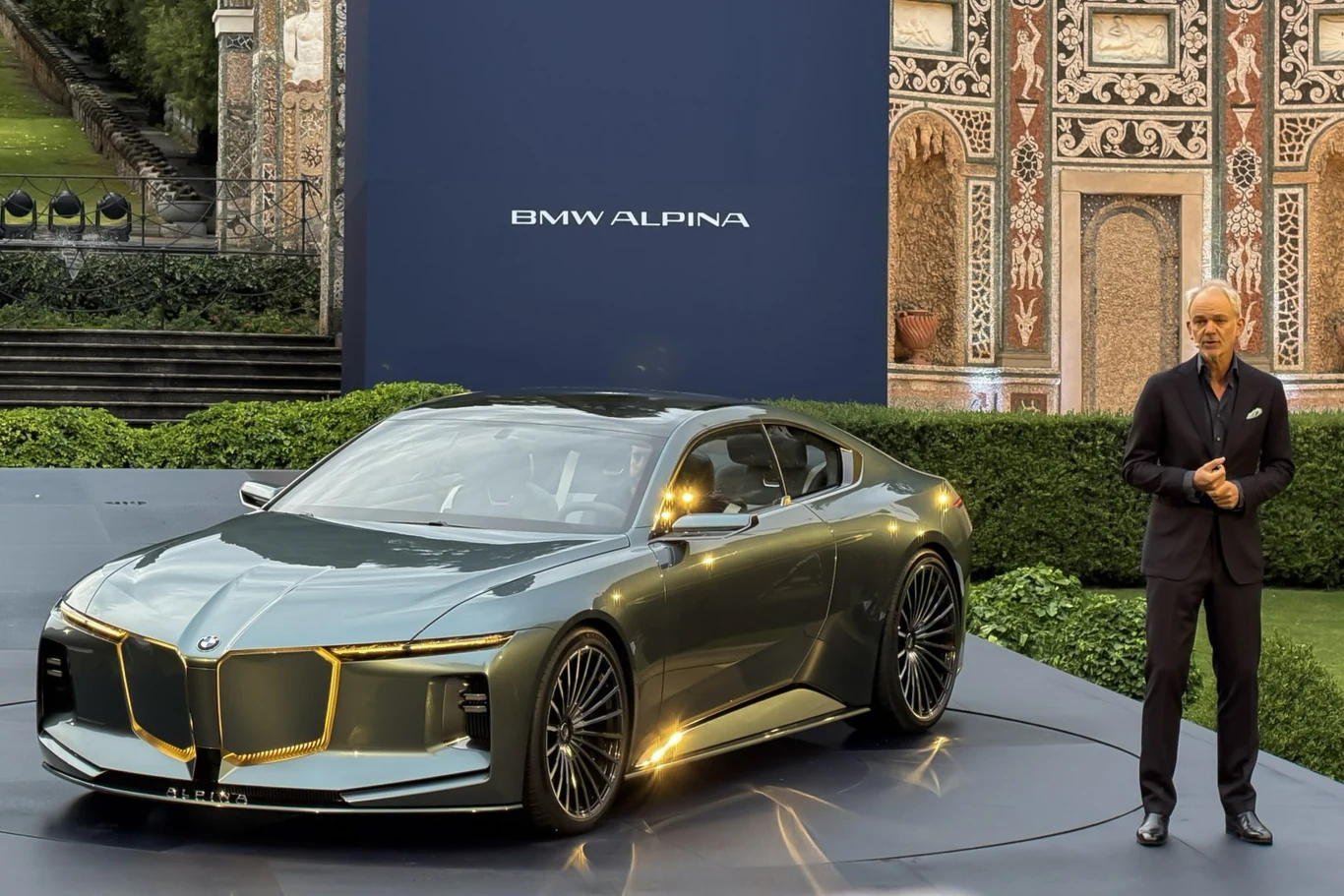 Adrian van Hooydonk powierzył misję stworzenia nowego modelu młodemu i zdolnemu projektantowi - Maximilianowi Missioniemu. Luksusowy samochód BMW Alpina w metalicznym odcieniu szarości z charakterystycznym, nowoczesnym designem stoi na podwyższonej platformie, obok mężczyzny w garniturze na tle eleganckiej architektury i plakatu z logo marki.