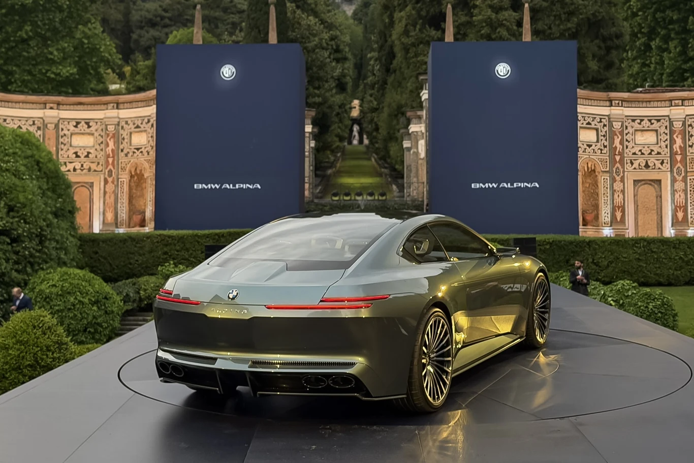 Największym zaskoczeniem w Vision BMW Alpina jest spalinowy silnik V8. Wszyscy spodziewali się elektrycznego napędu. Nowoczesny samochód sportowy BMW Alpina o opływowej sylwetce, ustawiony na podwyższonej platformie, otoczony zielenią i dekoracyjnymi panelami z logo marki w tle.