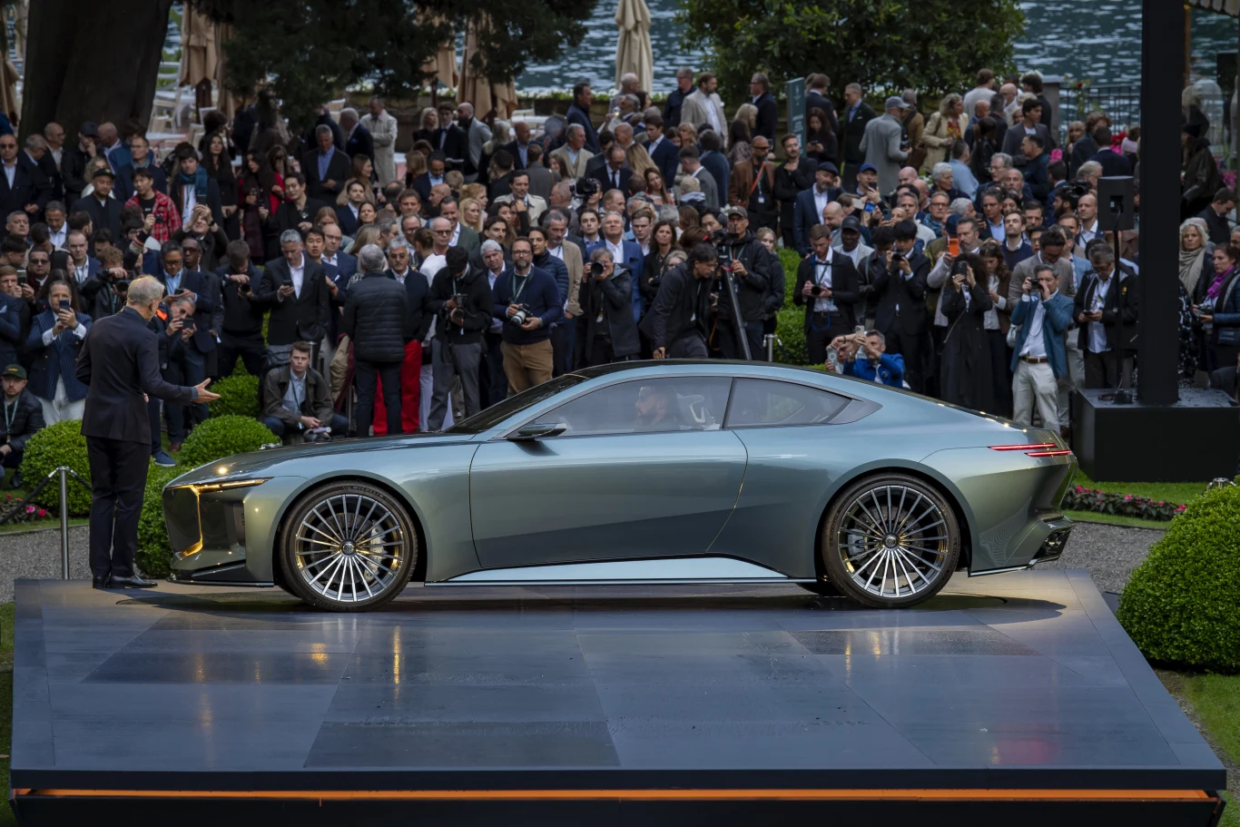 Vision BMW Alpina to pierwszy owoc połączenia niewielkiej manufaktury z Buchloe z potęznym koncernem. Srebrny samochód sportowy na podwyższonej platformie, otoczony przez tłum elegancko ubranych osób obserwujących prezentację.