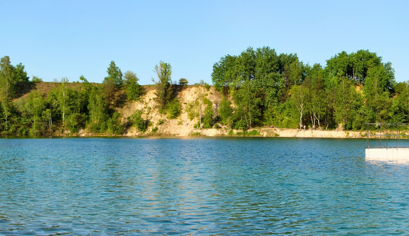 Gdzie jest "polski Balaton"? Błękitna woda otoczona stromym brzegiem porośniętym zielonymi drzewami, z fragmentem betonowego pomostu po prawej stronie.