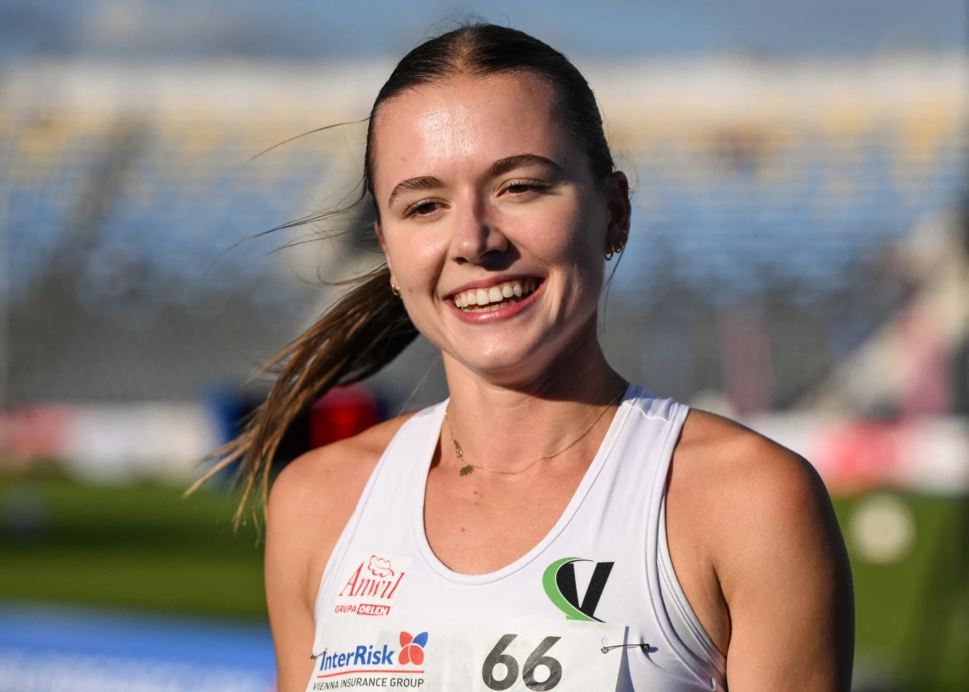 Klaudia Kazimierska Uśmiechnięta młoda kobieta w sportowym stroju z numerem startowym 66, z widocznymi logotypami sponsorów na tle rozmytego stadionu lekkoatletycznego.