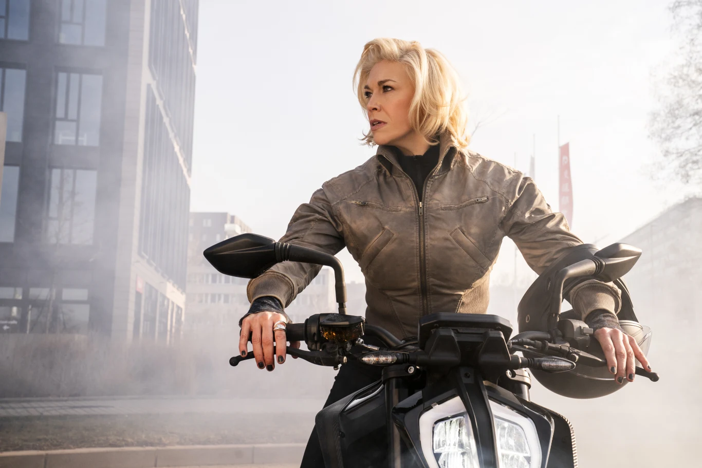 Hannah Waddingham w serialu "Kumpele na zabój" Kobieta o blond włosach w skórzanej kurtce siedzi na motocyklu, patrząc w bok z wyrazem skupienia na twarzy.