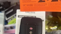 Czarny pas bagażowy z zamkiem szyfrowym oraz uchwytem w odcieniach zieleni i czerni, trzymany w lewej dłoni nad opakowaniem produktu z napisem Smukee Home.