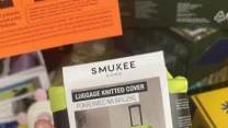 Ręka trzyma opakowanie jasnozielonego pokrowca na walizkę marki Smukee Home z widoczną etykietą produktu i zdjęciem pokrowca na walizce.