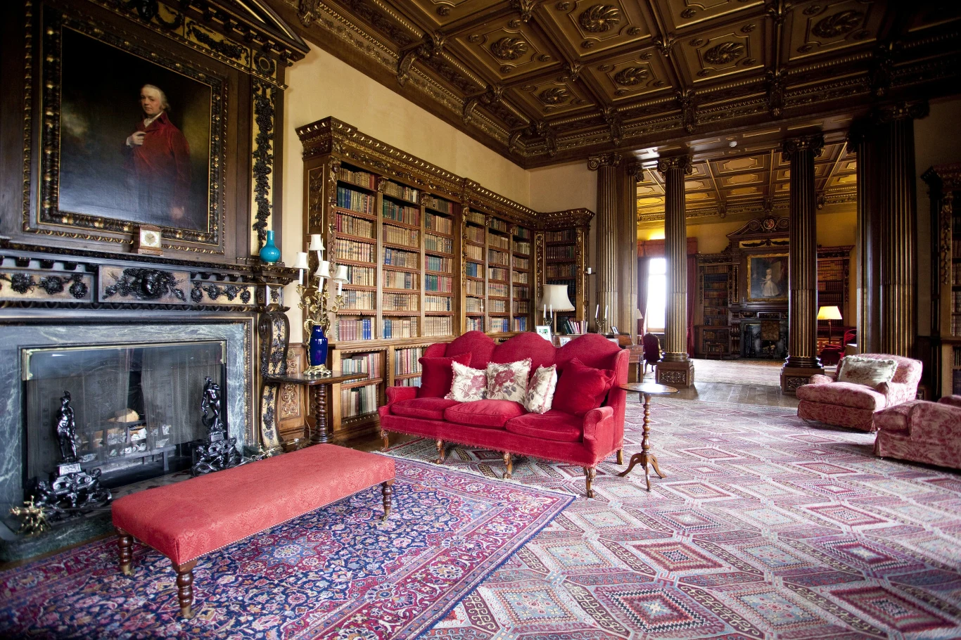 Bogato zdobiona biblioteka z wysokimi regałami pełnymi książek, ciemnym kominkiem, masywnym portretem w złotej ramie nad kominkiem oraz eleganckimi, bordowymi sofami i ławką ustawionymi na wzorzystym dywanie.