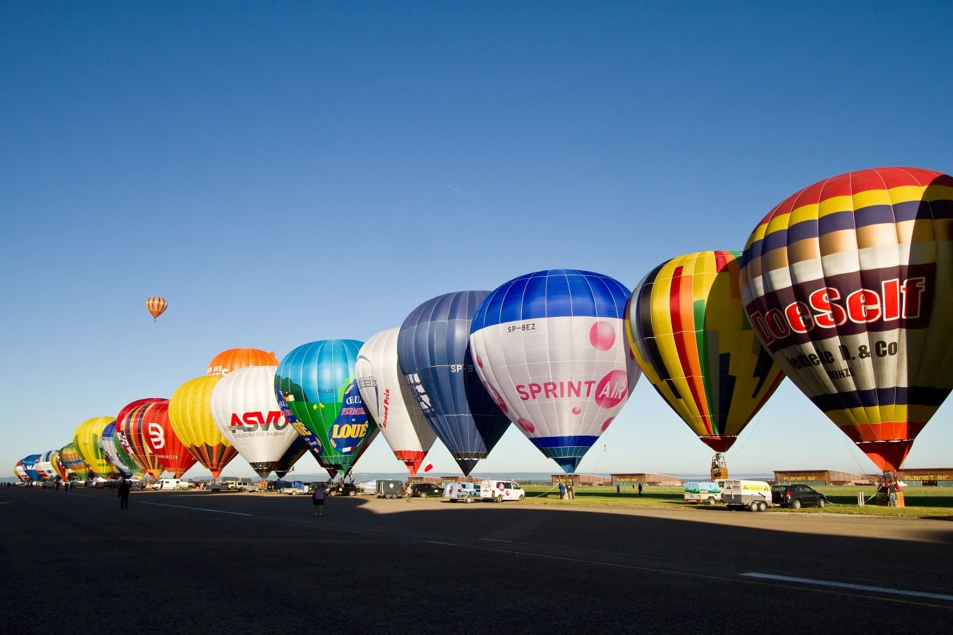 Kolorowe balony na gorące powietrze ułożone w szeregu na otwartym terenie, gotowe do startu o poranku pod błękitnym niebem.
