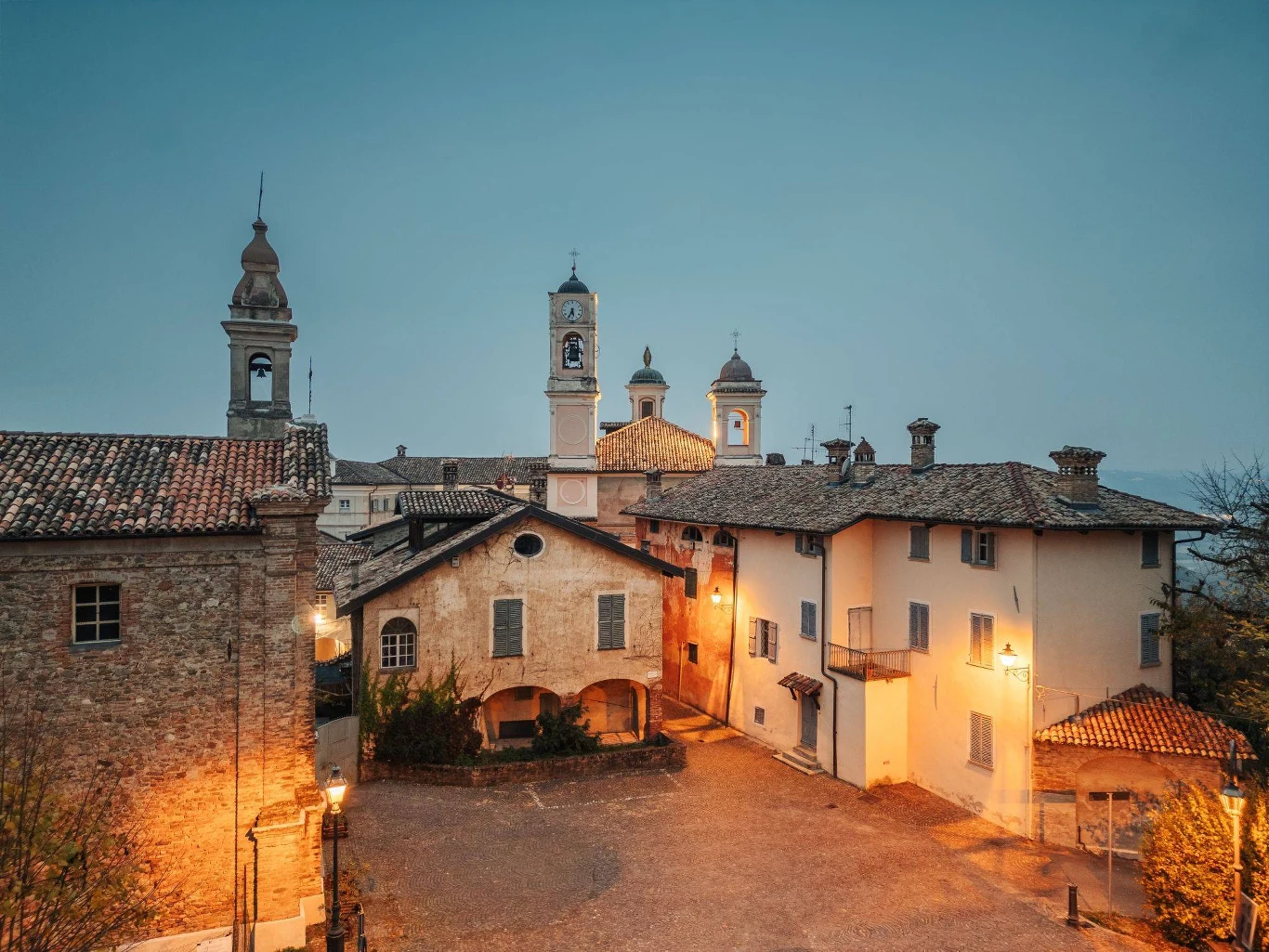 Borgo Monchiero Heritage - dawny klasztor i historyczna osada Kamienny dziedziniec otoczony zabytkowymi budynkami o czerwonych dachach, w tle widoczne wieże kościołów z dzwonnicami, podświetlone ciepłym światłem latarni o zmierzchu.