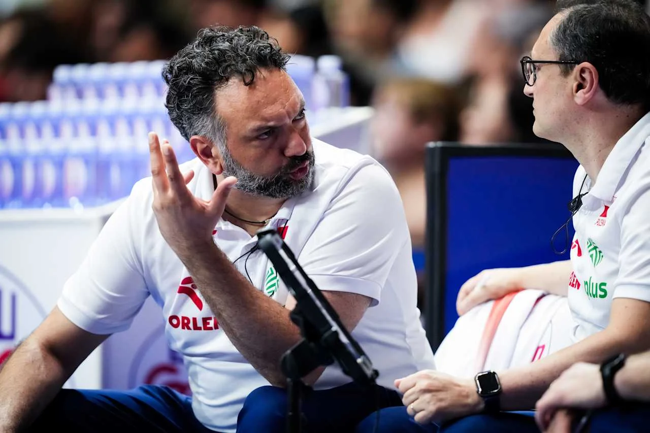Stefano Lavarini Dwóch trenerów w białych koszulkach z logo Orlen prowadzi ożywioną rozmowę podczas meczu siatkówki, jeden z nich gestykuluje dłonią, a w tle widoczne są rzędy butelek z wodą.
