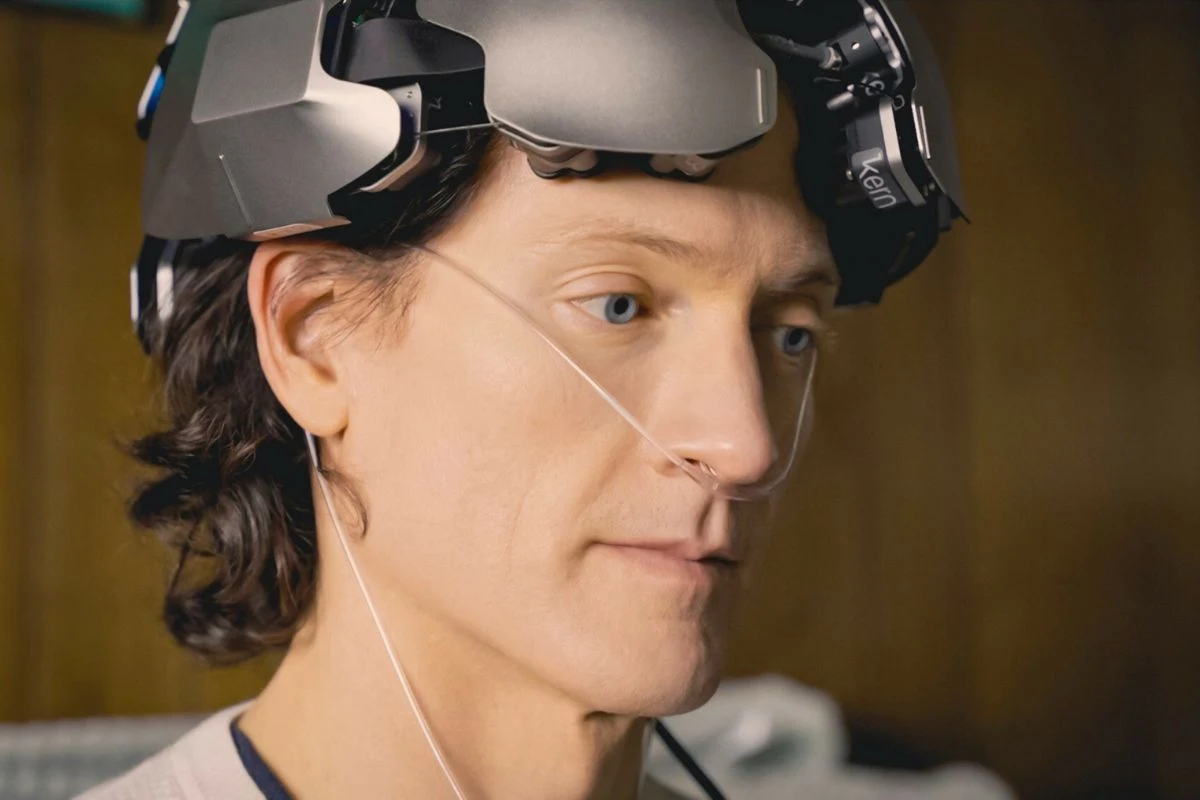 Mężczyzna z długimi włosami ma na głowie zaawansowane technologicznie urządzenie oraz założoną plastikową rurkę do nosa, co sugeruje udział w medycznych lub naukowych badaniach.