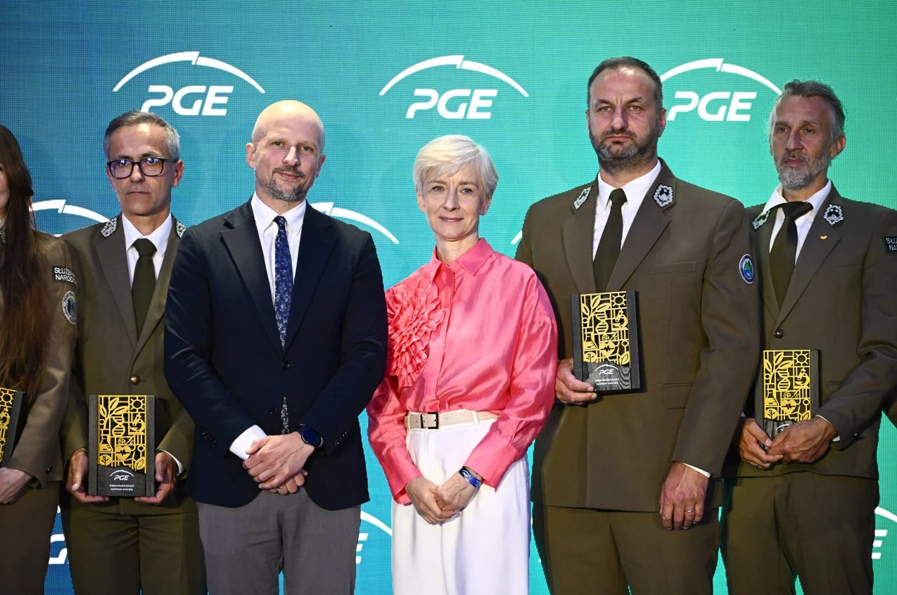Grupa uhonorowanych osób w eleganckich strojach, w tym mężczyźni w mundurach oraz kobieta w różowej bluzce, trzymająca w rękach nagrody na tle charakterystycznego zielonego motywu z logo PGE.