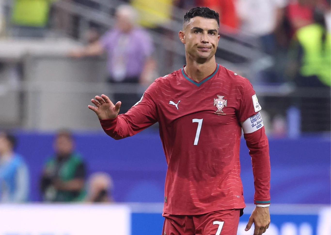 Cristiano Ronaldo Piłkarz w czerwonym stroju z numerem 7 i opaską kapitana na ramieniu unosi jedną rękę, stojąc na murawie stadionu sportowego.