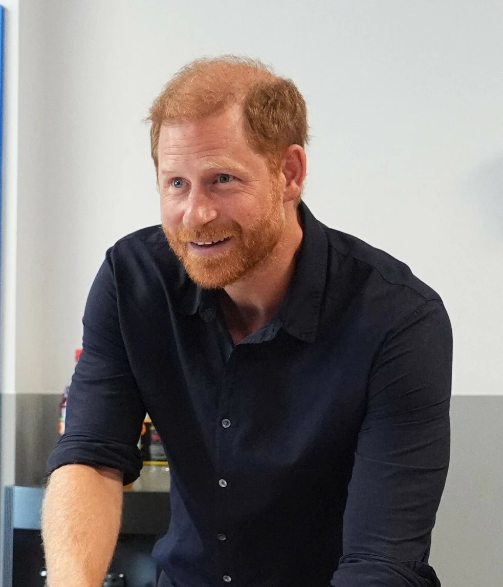 Książę Harry podczas pobytu w Anglii 9 lipca 2026 roku Mężczyzna o rudych włosach i brodzie, ubrany w ciemnoniebieską koszulę z podwiniętymi rękawami, pochyla się do przodu i uśmiecha.
